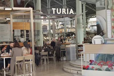 People dining at tables under a 'TURIA' sign in a market setting.