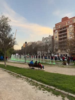 People sitting on a bench near a fountain in a park with buildings in the background.