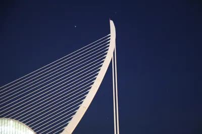 Close-up of a modern cable-stayed bridge with visible cables and a curved tower against a dark sky.