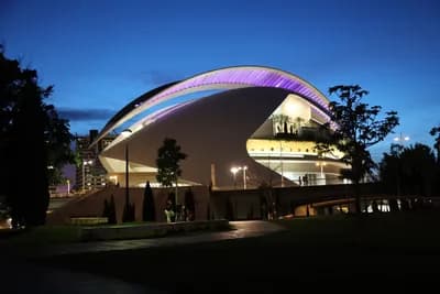 Modern architecture with curved roof illuminated at dusk, surrounded by trees and a clear sky.
