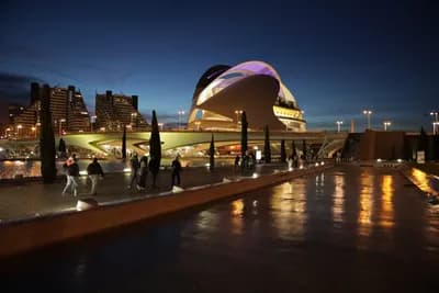 A modern, illuminated building with a unique, curved design reflected in a wet surface at night.