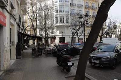 A street view with parked vehicles, a scooter, and café seating in front of a building.
