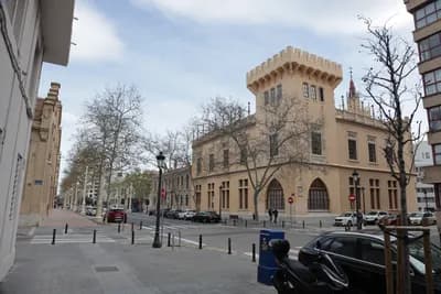 Historic building with arched windows and tower, parked cars, bare trees, cloudy sky.