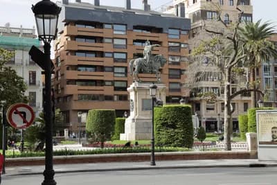 Statue of a person on horseback in a landscaped plaza with modern buildings and trees.