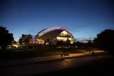 A modern building with a curved roof illuminated at dusk in a park setting.