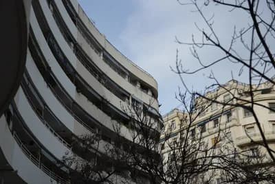 A curved white building with balconies, bare tree branches, and a clear blue sky.