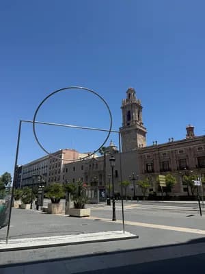 Modern art installation with a large circular sculpture in a city square with historic buildings and a clear blue sky.