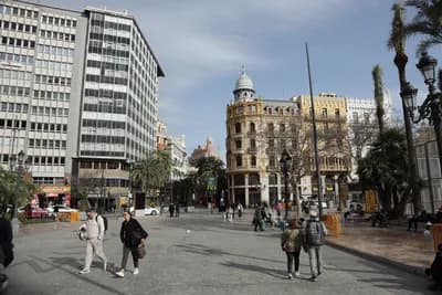 Pedestrians walking in a plaza with palm trees and historic buildings under a clear sky.