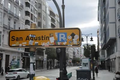 Straßenschild für San Agustín mit Parksymbol und Aufklebern, städtische Gebäude, bewölkter Himmel.