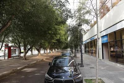 A street scene with parked cars, trees, and a modern building with a glass facade.