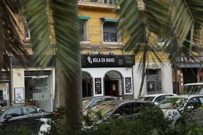 A yellow building with 'BOLA EN MANO' sign, palm fronds, and parked cars.