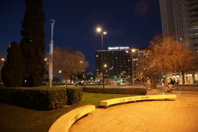A nighttime city square with a curved bench and a lit hotel in the background.