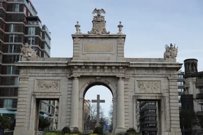 A historic stone archway with a cross, surrounded by modern urban architecture and a clear sky.