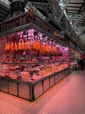 Market stall with hanging cured meats and food items under a metal archway.