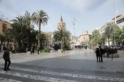 A plaza with palm trees, a church tower, and people walking in Valencia, Spain.