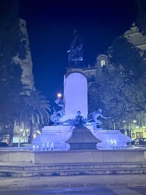 Statue illuminée la nuit avec des arbres et des bâtiments en arrière-plan.