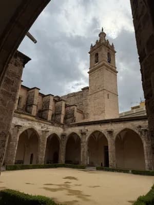 Cortile in pietra con passaggi ad arco e alto campanile sotto un cielo nuvoloso.