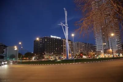 A nighttime cityscape featuring illuminated buildings and a tall sculpture.