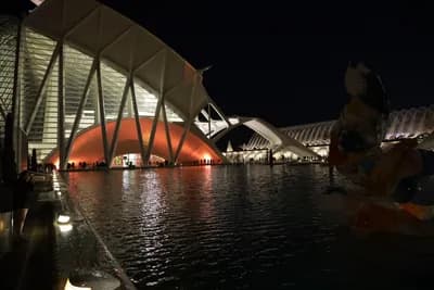 A person in a costume sitting on the ground in front of a modern, illuminated architectural structure at night.