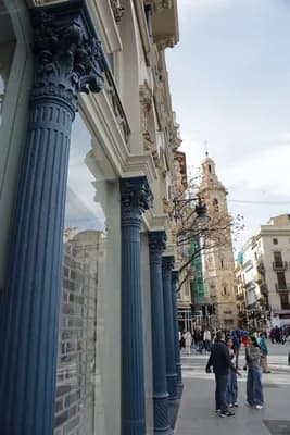 Ornate blue columns line a street in Valencia, Spain, with people walking and a church tower in the background.
