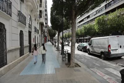 Two people walking on a sidewalk lined with trees and parked vehicles.