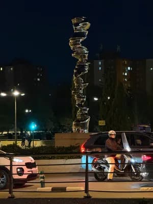 A nighttime scene featuring a person on a scooter near an abstract sculpture and parked cars.