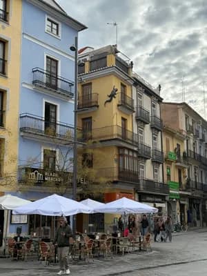 Street scene with colorful buildings, outdoor cafes, and pedestrians under a cloudy sky.