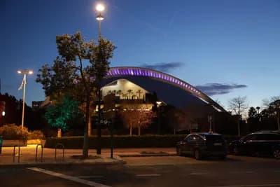 A modern, illuminated building at dusk with trees and parked cars in the foreground.