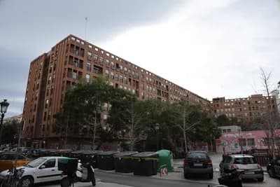 Urban street with brick building, parked cars, and trash bins under a cloudy sky.