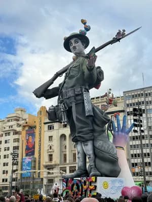 A large statue of a soldier holding a rifle in a public square with buildings and a crowd in the background.