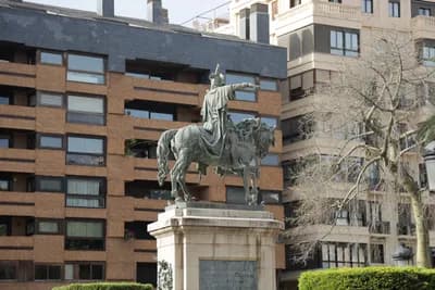 Statue of a person on horseback in front of a modern building with a tree in the foreground.