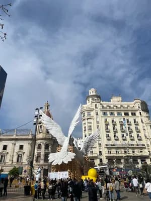 A large white dove sculpture in a plaza with historic buildings and people gathered in the foreground.