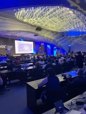 A conference audience in a large auditorium with a speaker on stage and a projection screen.