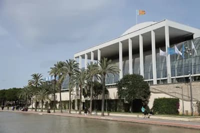 Modern building with palm trees and a flag, near a waterfront promenade with people walking.