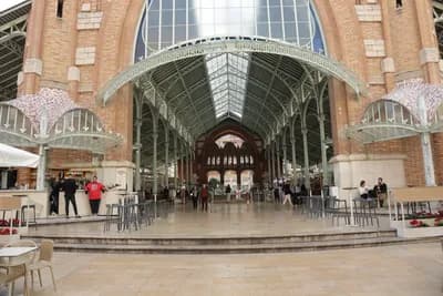 A large arched entrance with glass roof and brick walls, featuring a bustling interior with people and tables.