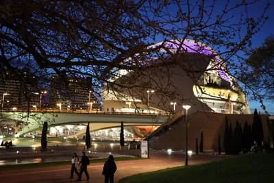 Modern building with curved roof at dusk, people walking, bridge in background.