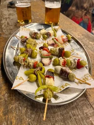 Skewered appetizers with olives and peppers on a tray, accompanied by two glasses of beer.