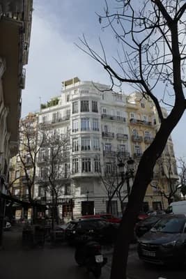 A white building with balconies and a tree in the foreground on a street.