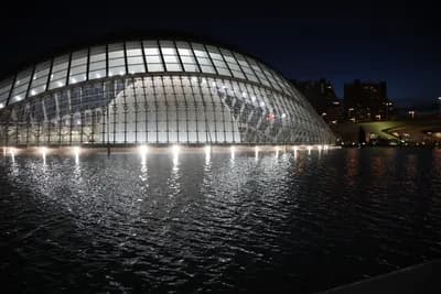A modern, dome-shaped building illuminated at night, reflected in calm water.