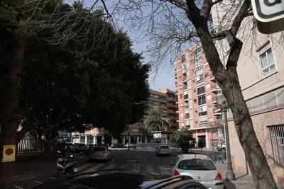 Urban street with parked cars, trees, and a multi-story building under clear skies.