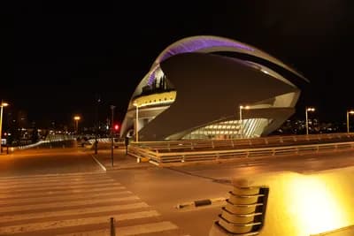 A modern, illuminated building with a curved design at night, surrounded by a pedestrian walkway and streetlights.