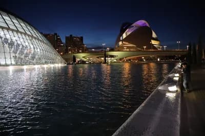 A nighttime view of a futuristic building with a person walking along a waterfront promenade.