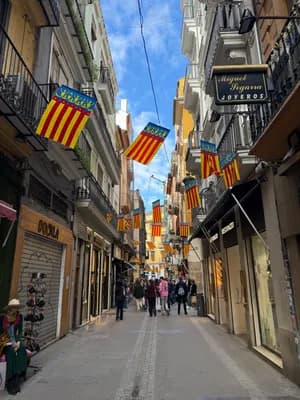 A street scene with Catalan flags and shops, clear blue sky above.