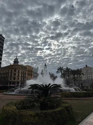 A fountain with water jets surrounded by palm trees and historic buildings under a cloudy sky.