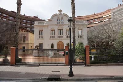 A white building with ornate architecture and a metal gate in front, surrounded by trees and other buildings.