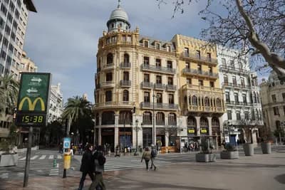 Historic building with ornate architecture in a plaza with pedestrians and a digital clock.