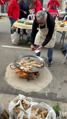 A man cooking paella over an open fire on a street with people in the background.
